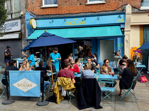 People dining outside The Sicilian Deli on a sunny day near Brockwell Park